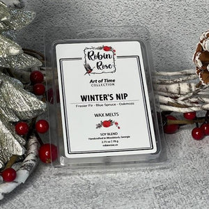 Wax melt container standing up next to a christmas tree. Background has berries and pine combs. Label on container has a robin and a rose on it.