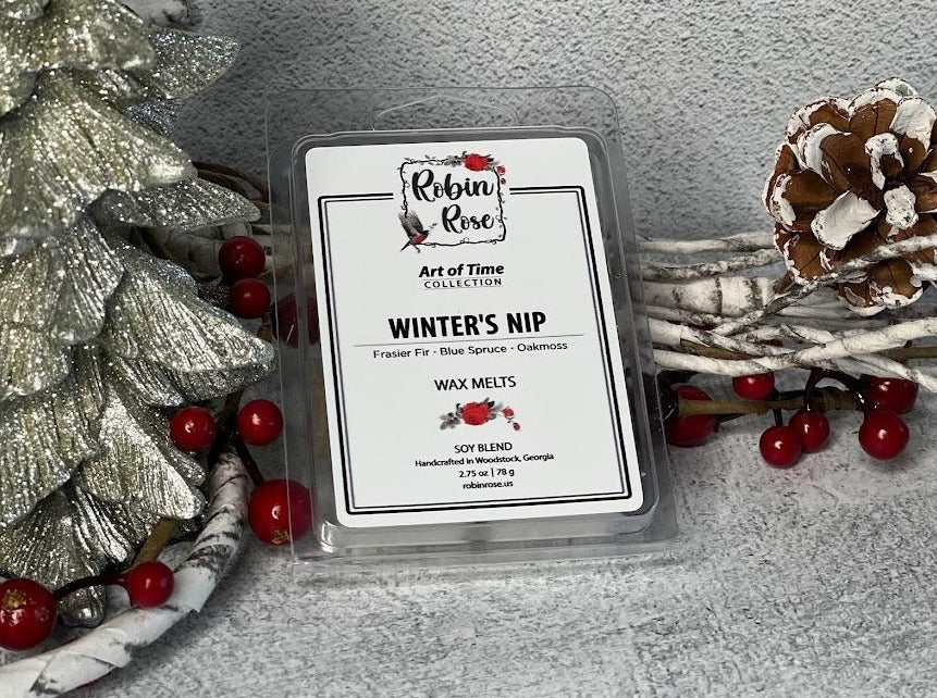 Wax melt container standing up next to a christmas tree. Background has berries and pine combs. Label on container has a robin and a rose on it.